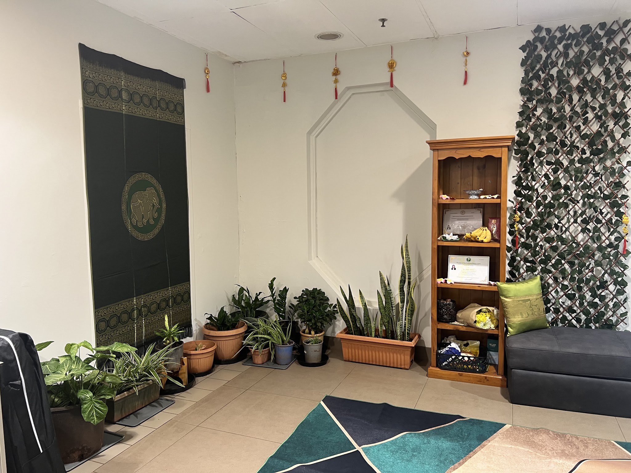 The waiting area at Newman Massage, with potted plants, a green and gold Thai wall hanging and a shelf of ornaments